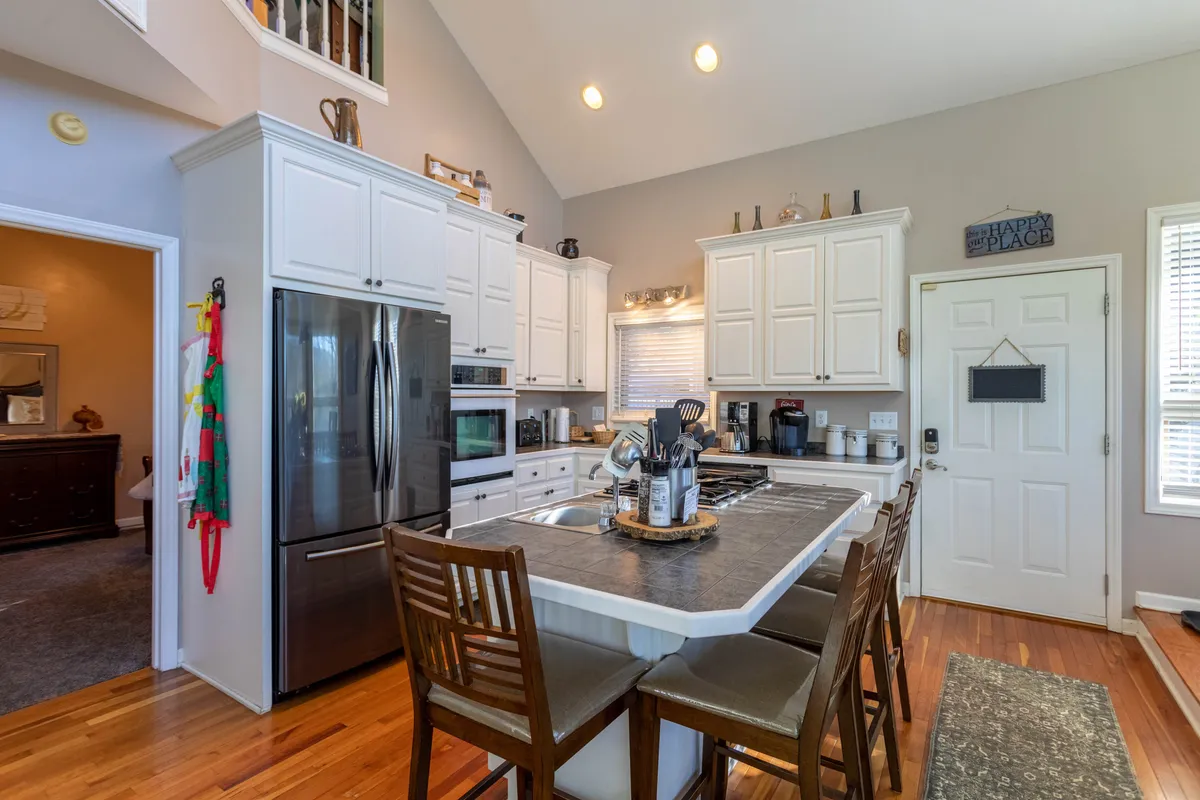 This image showcases a bright, inviting kitchen with an island countertop, stainless steel appliances, and wooden chairs. The kitchen is styled for gatherings, featuring decorative items on shelves and an open space ideal for cooking and entertaining.