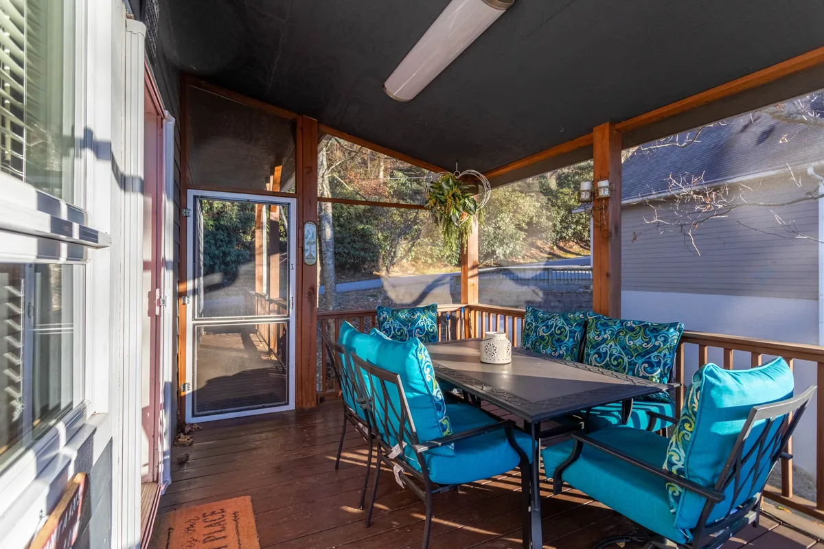 A cheerful screened-in porch featuring a dining table with colorful cushions and visible porch structure. The space is flooded with natural light and surrounded by greenery, creating an inviting spot for outdoor dining, morning coffee, or evening gatherings.