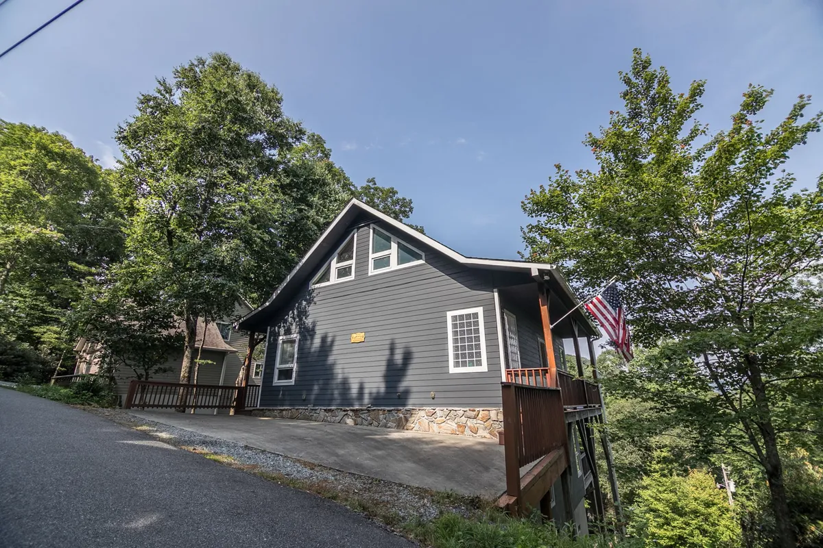 This image presents the exterior of Misty Mountain Hideaway, featuring the blue house with an American flag and surrounded by tall, leafy trees. The spacious porch invites relaxation and gatherings, while the lush natural setting hints at the proximity to outdoor adventures and family-friendly activities.