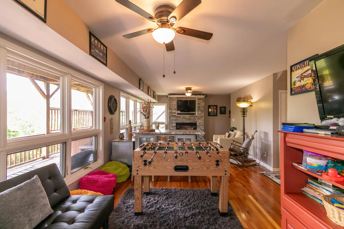 This image showcases a lively lower-level living space with a foosball table, sofa, fireplace, TV, and large windows bringing in light. The setting is casual and perfect for playing games, relaxing, or watching movies together.