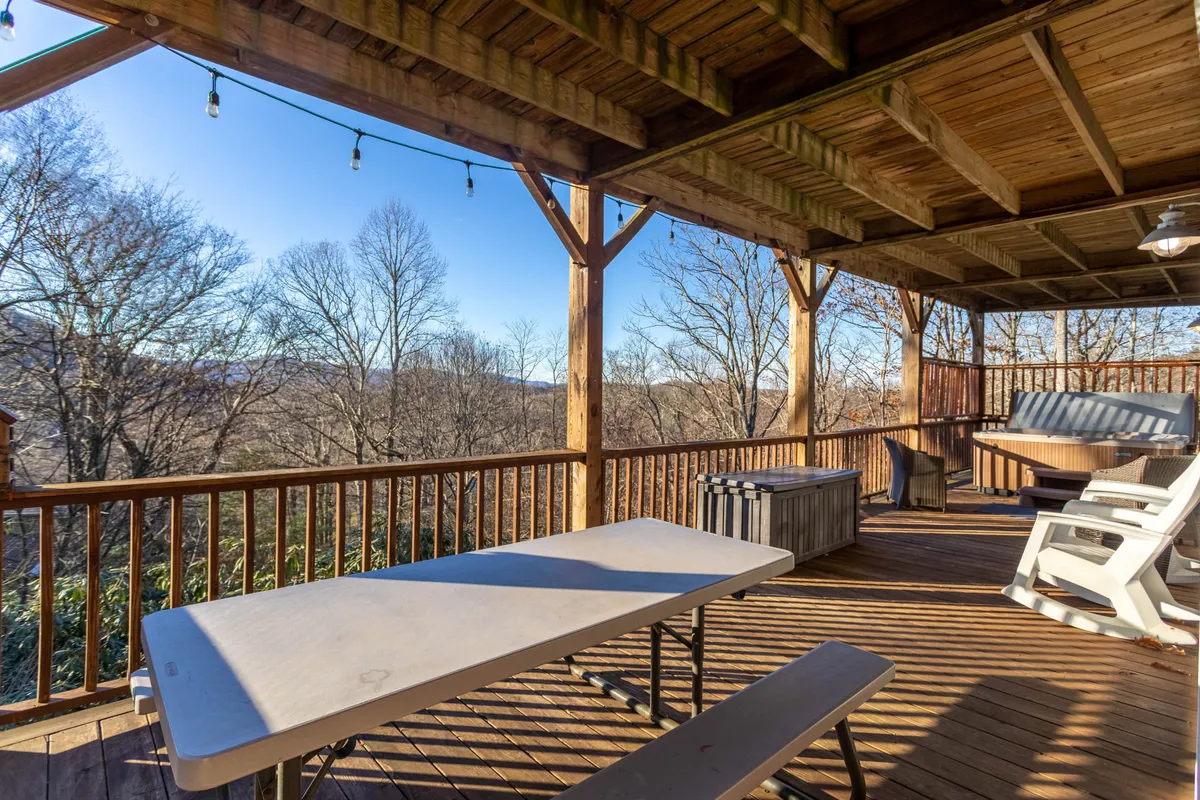 An exterior shot showing a spacious wooden deck furnished for outdoor dining, surrounded by trees and offering mountain views. The setting suggests relaxed gatherings, sunset views, and outdoor entertainment options.