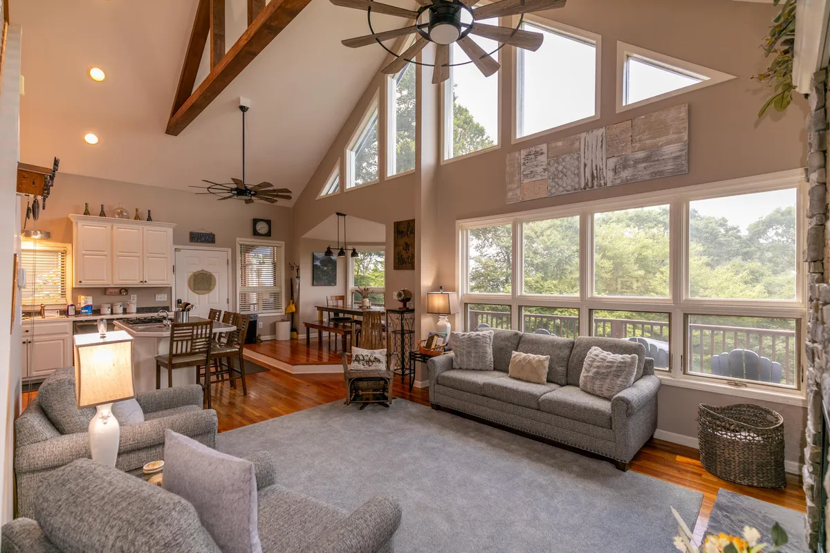 A spacious, open-plan living area with large windows reveals sweeping mountain views. Plush sofas are arranged for comfort, with a ceiling fan and exposed wooden beams adding to the mountain lodge ambiance. The dining area, visible in the background, is set beneath a picturesque bay window, offering a seamless flow between spaces.