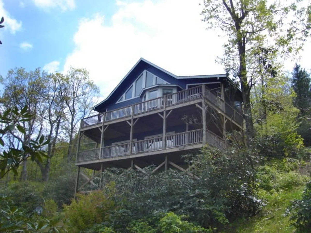 A charming mountain home nestled among lush greenery, featuring an expansive deck perfect for relaxing or gathering with friends and family. The peaceful setting showcases sunset views over the treetops, with plenty of space to enjoy stargazing or morning coffee surrounded by nature.