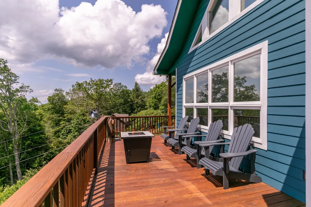 This image displays the exterior of the blue house with deck chairs arranged around a fire pit, surrounded by lush greenery. The setting is serene and welcoming, with plenty of outdoor space visible—the deck and the fenced area are especially prominent.